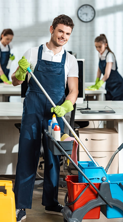 A smiling man in work attire holds a cleaning pole in an office setting, surrounded by colleagues in the background. Cleaning supplies are organized in a cart nearby.