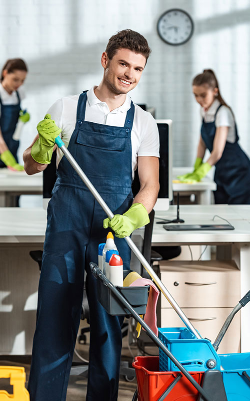 A smiling male window cleaner in a blue uniform holds a cleaning tool and is surrounded by colleagues working in an office setting. Bright light streams in through the windows, highlighting the clean environment.