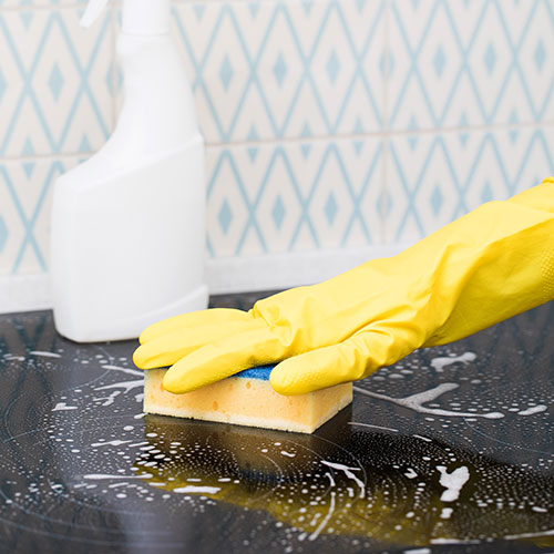 A person wearing yellow gloves scrubs a surface with a sponge, surrounded by cleaning supplies. The background features a patterned wall, suggesting a cleaning environment.